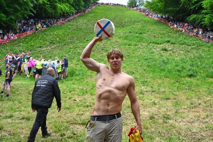 Young man holding a round object on a grassy hill during a strange tradition event with a crowd watching from the sides