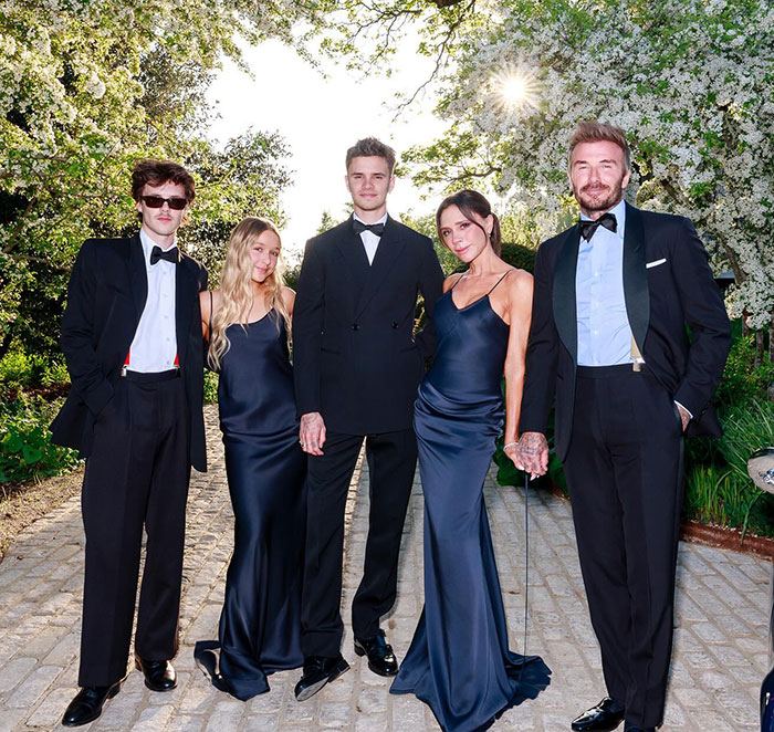 Nicola Peltz with tattoo and family members dressed in formal attire outdoors near blossoming trees at sunset.