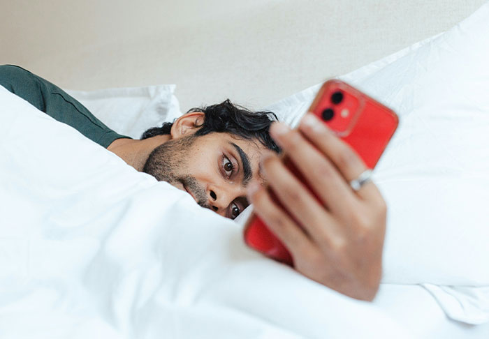 Man lying in bed using a smartphone, focused on improving life skills to retire rich and achieve financial success.