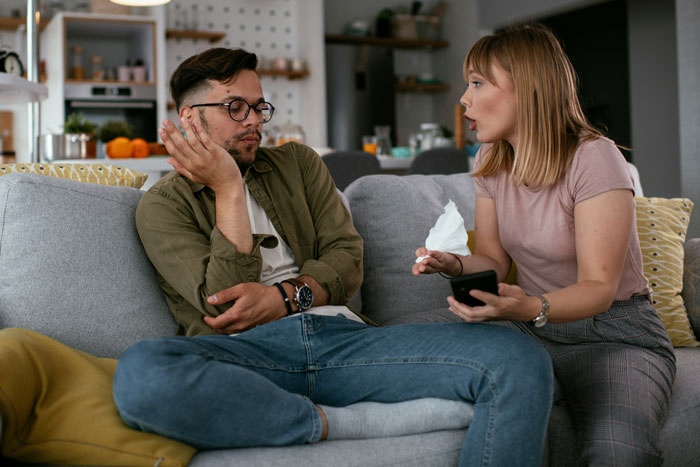 Unemployed guy on couch acting dismissive as friend holds phone and tissue, arguing in a living room