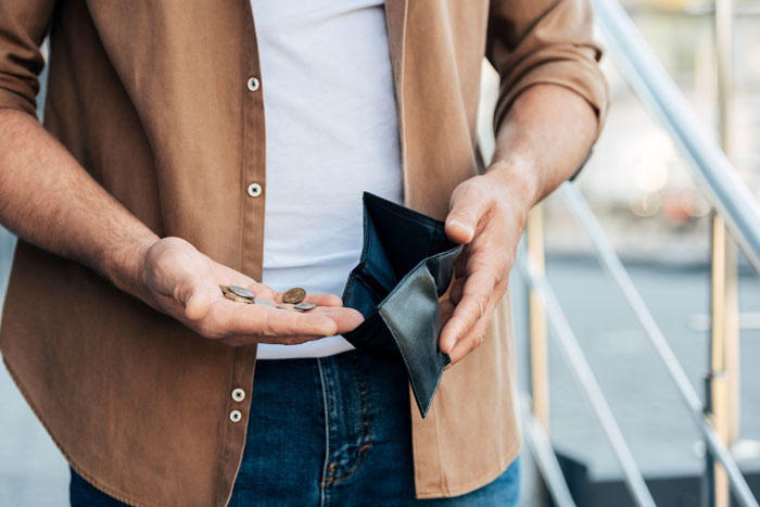 Unemployed guy holding empty wallet and loose coins, wearing brown shirt and jeans on outdoor steps