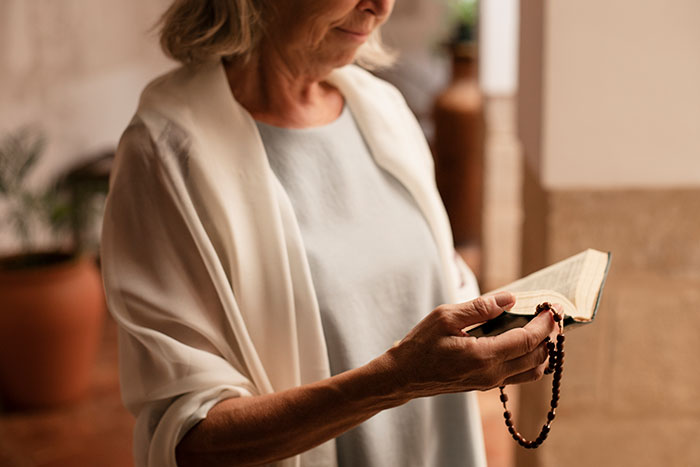 Religious lady holding rosary and Bible, deeply focused on prayer in a peaceful indoor setting.