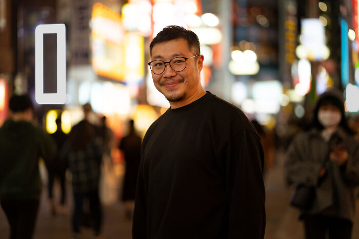Smiling bilingual man wearing glasses stands on a busy city street at night where gossip might happen.