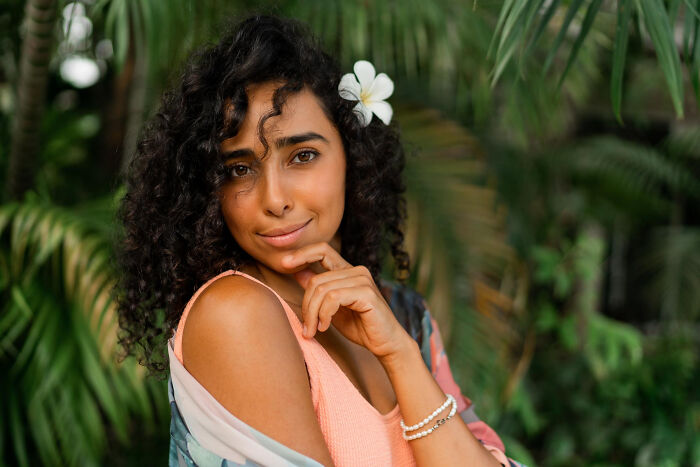 Young bilingual woman with curly hair and flower in hair, smirking as if caught someone gossiping about her outdoors.