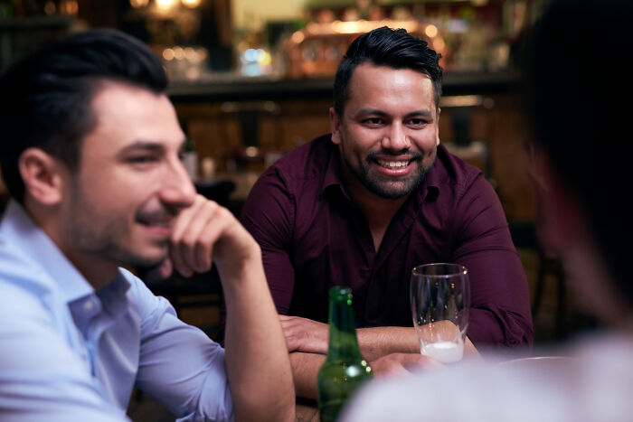 Three men talking and laughing at a bar, illustrating bilingual people catching someone gossiping about them.