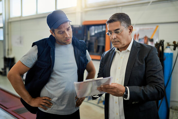 Two men in a workshop, one in casual workwear and the other in a suit, discussing documents about bilingual gossip instances.