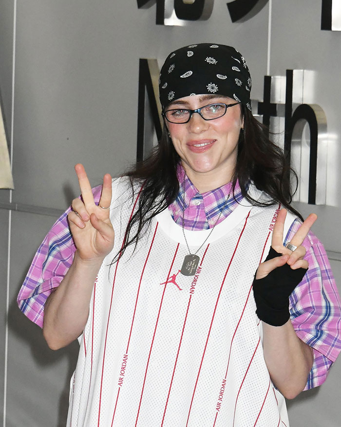 Billie Eilish wearing glasses and a bandana, smiling and flashing peace signs in a casual outfit.