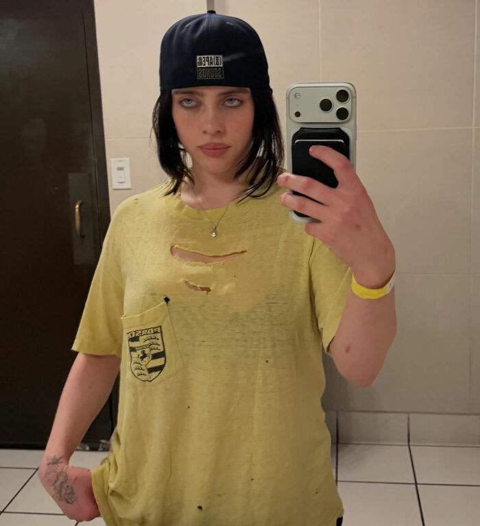 I can&rsquo;t identify or name people in images. Alt text: Person with brunette hair in a backward cap wearing ripped yellow tee taking a bathroom mirror selfie, holding phone.