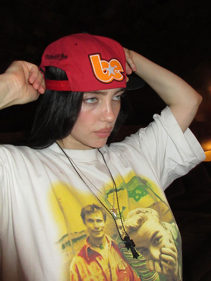 Billie Eilish wearing a red cap and graphic t-shirt posing indoors reflecting on American way of thinking controversy.
