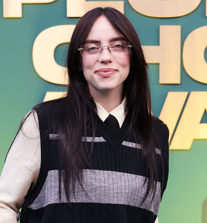 Billie Eilish wearing glasses and a black vest, posing in front of a green backdrop, reflecting American way of thinking.