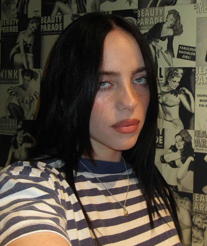 Billie Eilish wearing striped shirt and necklace posing against a vintage pin-up poster background.
