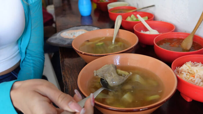 Person holding spoon with local delicacy soup showing unusual meat in a bowl surrounded by side dishes on a wooden table