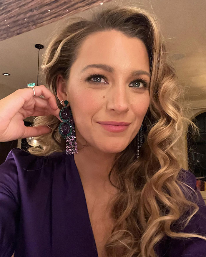 Blake Lively in a purple blouse, wearing large earrings, smiling in an indoor setting with soft lighting.
