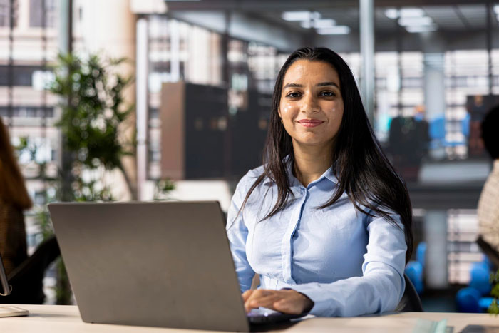 Young employee with South Asian ethnicity working on laptop in modern office, showing confident and hopeful expression. Young employee with South Asian ethnicity working on laptop in modern office, showing confident and hopeful expression.