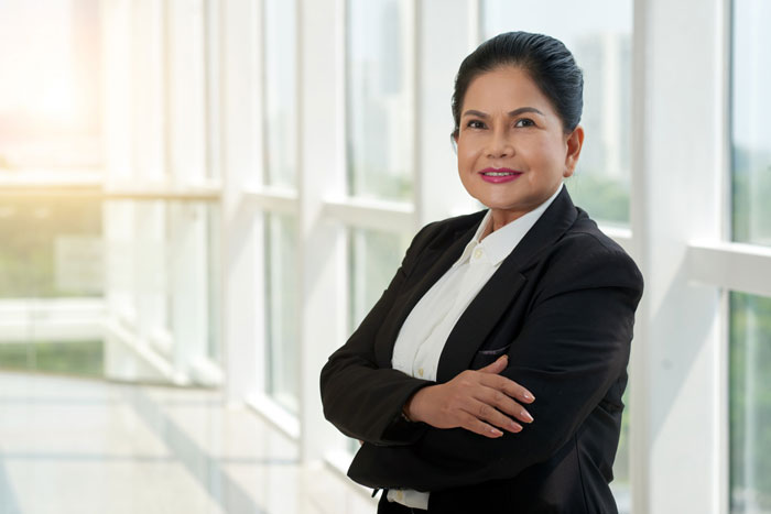 Confident businesswoman in a black blazer standing with arms crossed by office windows, depicting bullied employee concept. Confident businesswoman in a black blazer standing with arms crossed by office windows, depicting bullied employee concept.