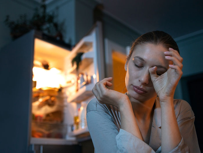 Woman sitting tired and stressed in front of an open fridge, triggered by childhood trauma during Christmas tradition.