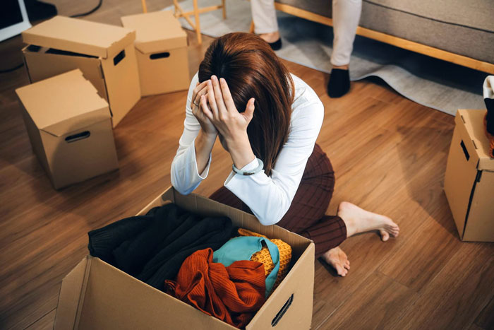 Woman feeling like a guest in her own home, sitting on floor overwhelmed while packing belongings in moving boxes.
