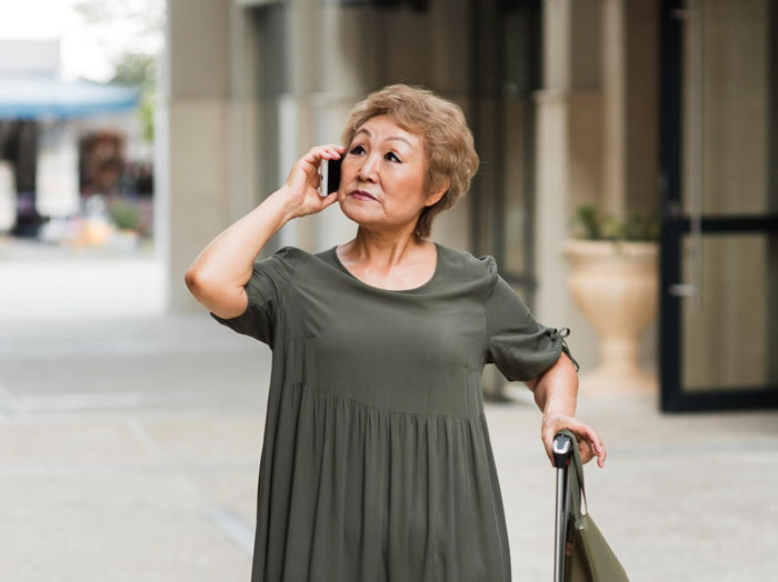 Older woman talking on phone outside building, representing feeling like a guest in her own home situation.