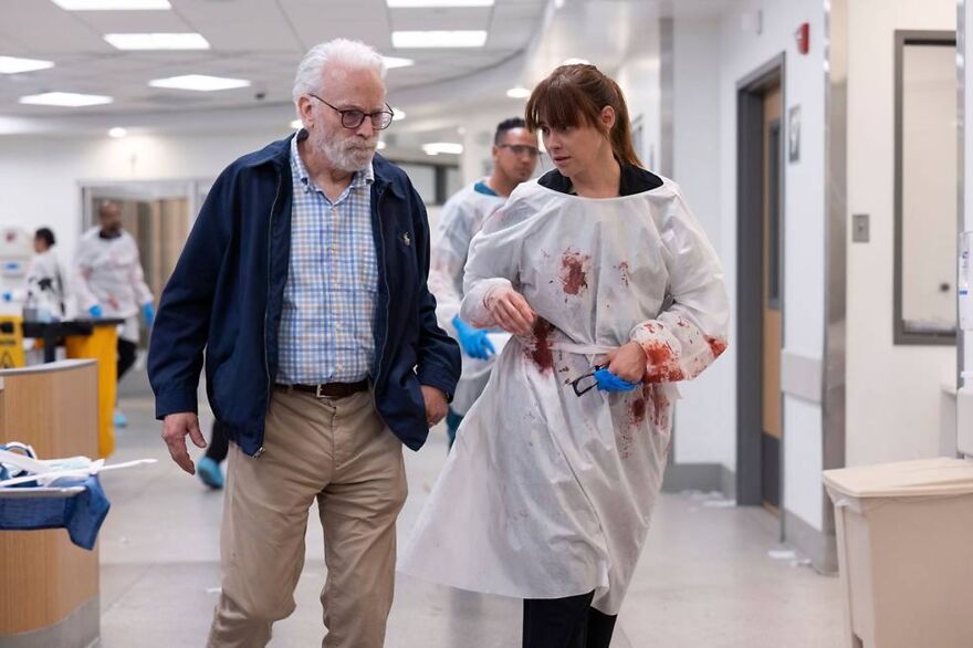 Two cast members of The Pitt walking in a hospital corridor, one wearing a blood-stained gown, discussing a scene.