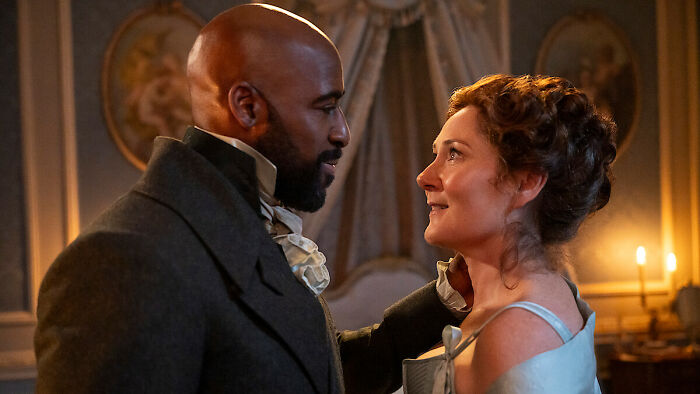 Two Bridgerton Season 4 cast members in period costumes sharing an intimate moment in a dimly lit room.