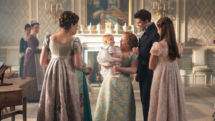Bridgerton Season 4 cast dressed in period costumes, interacting in an elegant room with a baby and vintage decor.