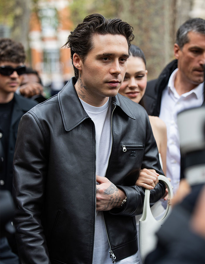 Brooklyn Beckham wearing a black leather jacket and white shirt, surrounded by people, in an outdoor setting.