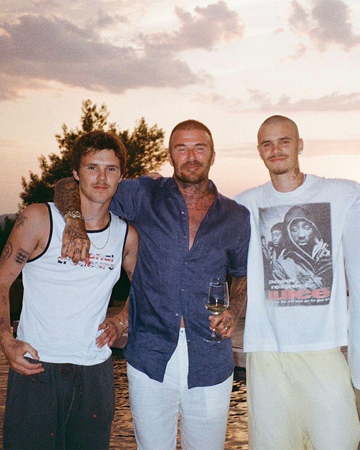 Brooklyn Beckham with family outdoors at sunset, casually dressed, with a glass of wine, amid family drama discussions.