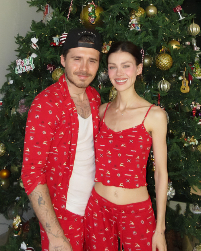 Brooklyn Beckham smiling with partner in matching red outfits standing in front of decorated Christmas tree during holiday season.