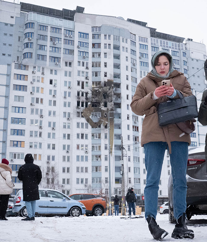 Woman in winter clothes using phone near damaged residential building in Ukraine amid tone deaf message backlash involving Timoth&eacute;e Chalamet fans.