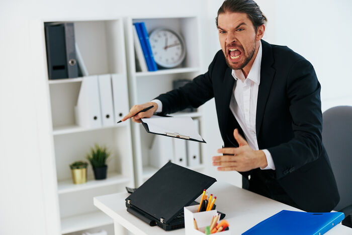 Angry male coworker in office throwing papers and acting unhinged showing signs of immediate firing risk.