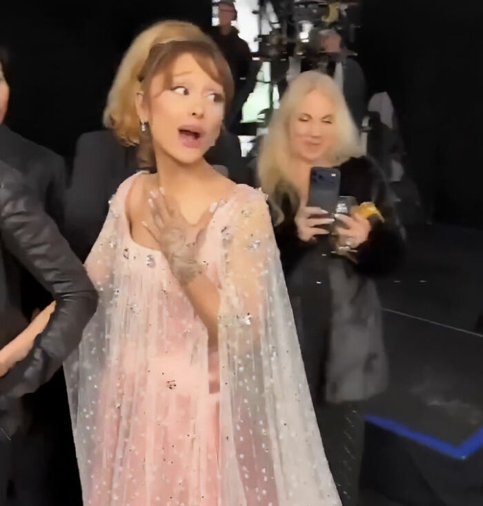 Ariana Grande reacting on the red carpet wearing a pink embellished dress with a sheer cape during an event. Ariana Grande reacting on the red carpet wearing a pink embellished dress with a sheer cape during an event.