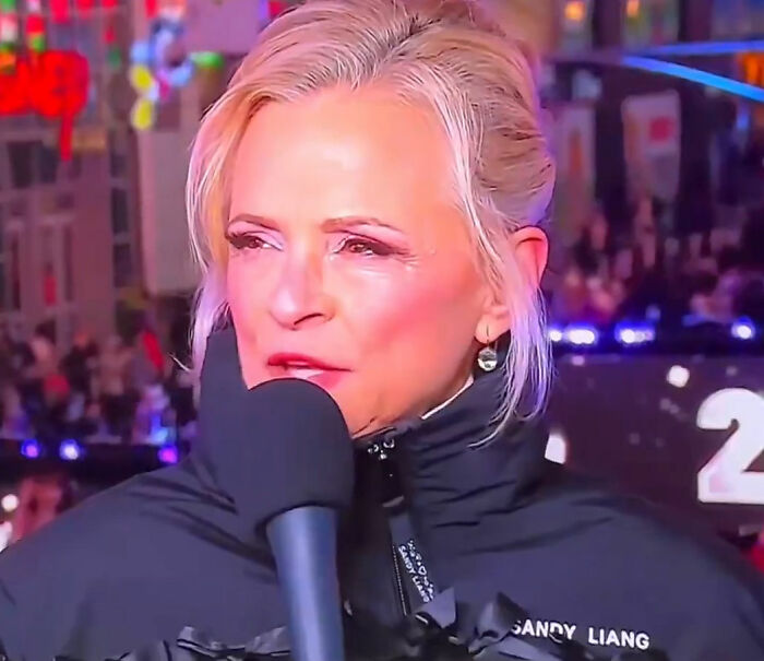 Amy Sedaris speaking into a microphone during CNN New Year's Eve broadcast with colorful crowd and lights in background.