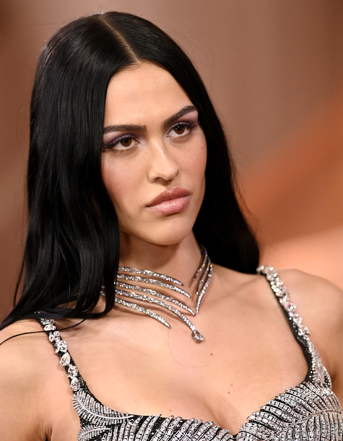 Woman with glowing skin and sleek black hair wearing a sparkling silver dress and statement necklace at a formal event.