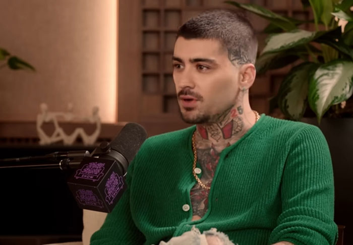 Man with tattoos wearing a green sweater speaking into a microphone in a casual podcast setting about celebrity infidelity scandals.
