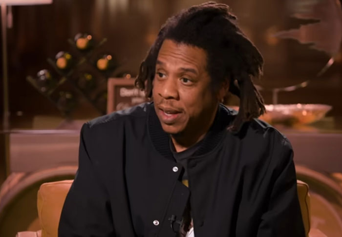 Man with dreadlocks wearing a black jacket in an indoor setting discussing celebrity infidelity scandals and relationships.