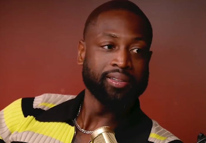 Man with beard and chain speaking into a microphone against a red background in a celebrity infidelity scandals context.