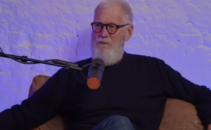 Older man with white beard and glasses speaking into a microphone during a discussion on celebrity infidelity scandals.