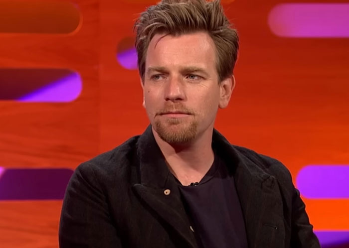 Actor sitting on a talk show set with red and purple background, related to celebrity infidelity scandals discussion.