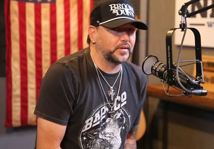 Man wearing a black cap and graphic t-shirt speaking into a microphone in a podcast studio about celebrity infidelity scandals.
