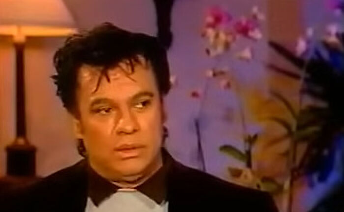 Man in a dark blazer during a celebrity interview, showing a thoughtful expression in a softly lit room with floral background.