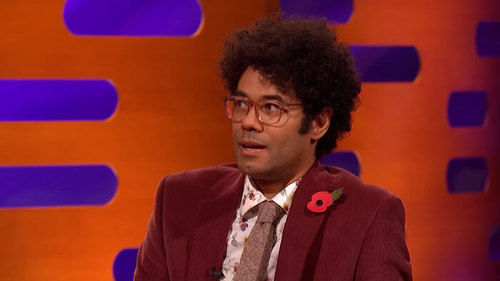 Man with glasses and curly hair in a burgundy suit giving a savage response during an interview on a talk show set.