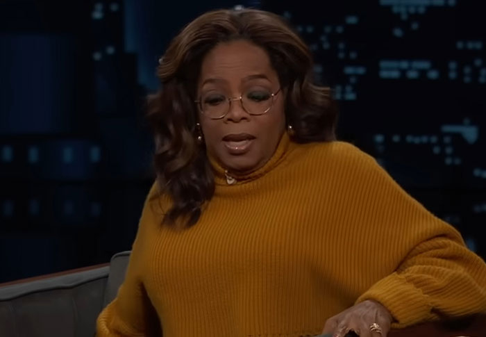 Celebrity in mustard sweater and glasses appearing out of touch with reality during a nighttime talk show interview.