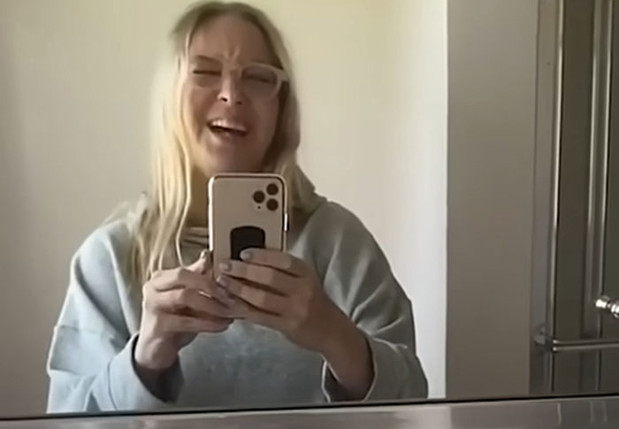 Celebrity taking a selfie in front of a mirror, laughing and wearing glasses and a grey sweatshirt, out of touch with reality.