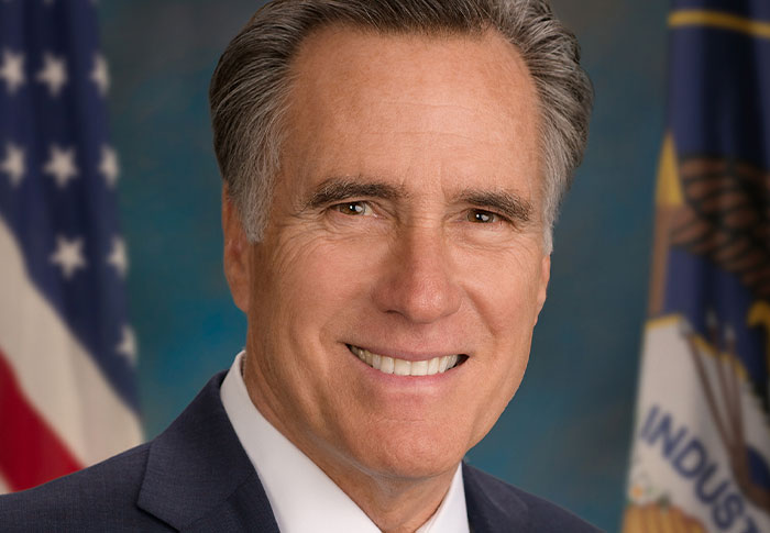 Portrait of a smiling man in a suit with American flags in the background, illustrating celebrities out of touch with reality.