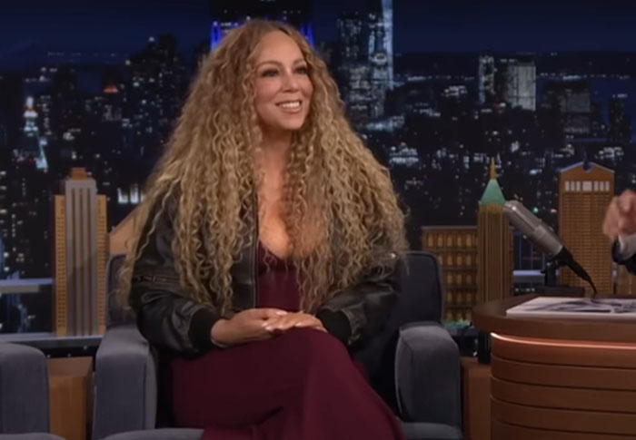 Celebrity with long curly hair wearing a black jacket and maroon dress during a late night talk show interview.