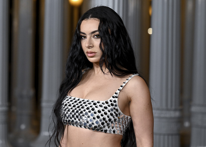 Charli XCX with long black hair wearing a silver studded braless top at an elegant columned venue.