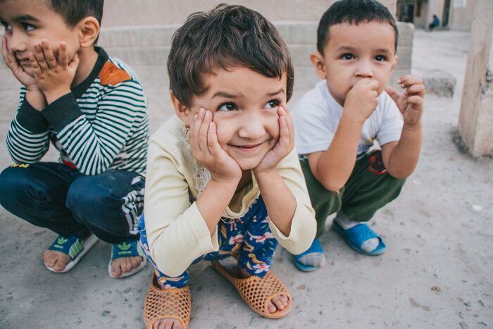 Three children squatting outdoors with playful expressions, illustrating cultural behaviors normal in their country but offensive elsewhere.