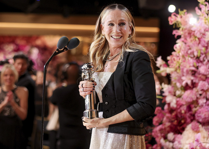Sarah Jessica Parker holding an award on stage, smiling during an event surrounded by pink floral decorations.