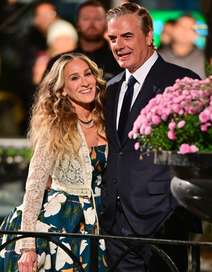 Chris Noth and Sarah Jessica Parker posing together at an event with flowers in the foreground and blurred crowd behind.