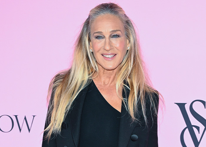 Sarah Jessica Parker posing at an event, wearing a black outfit with long blonde hair against a pink background.